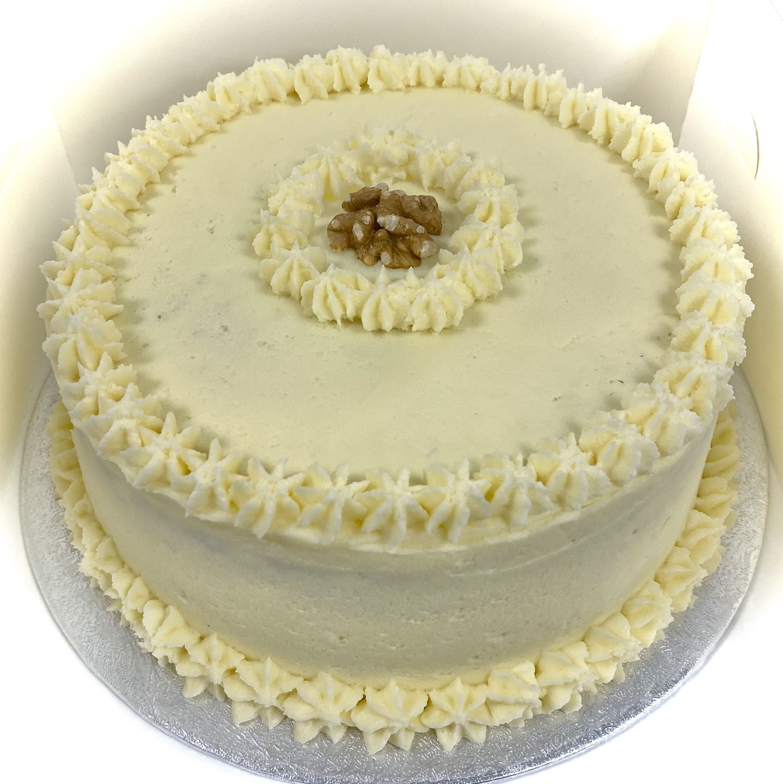 A round, award-winning carrot cake with smooth white frosting, decorated with two borders of small piped stars along the top edge and a single layer of piped stars in the center, where a small cluster of walnuts is placed. It is set on a silver cake board.